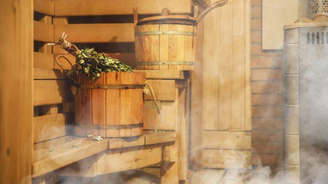 Lämmin sauna. Kiukaasta nousee hyöryä ja saunan lauteilla on saunavihta ja vettä täynnä oleva saunakiulu.